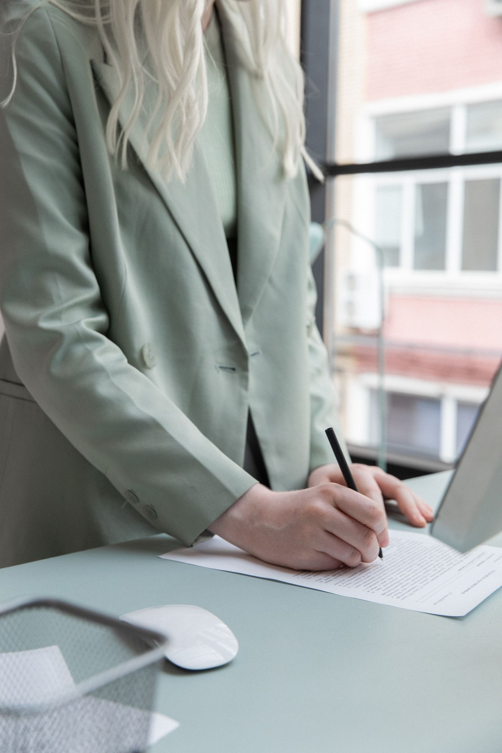 Mulher de blazer anota em um documento apoiado na mesa ao lado do notebook.