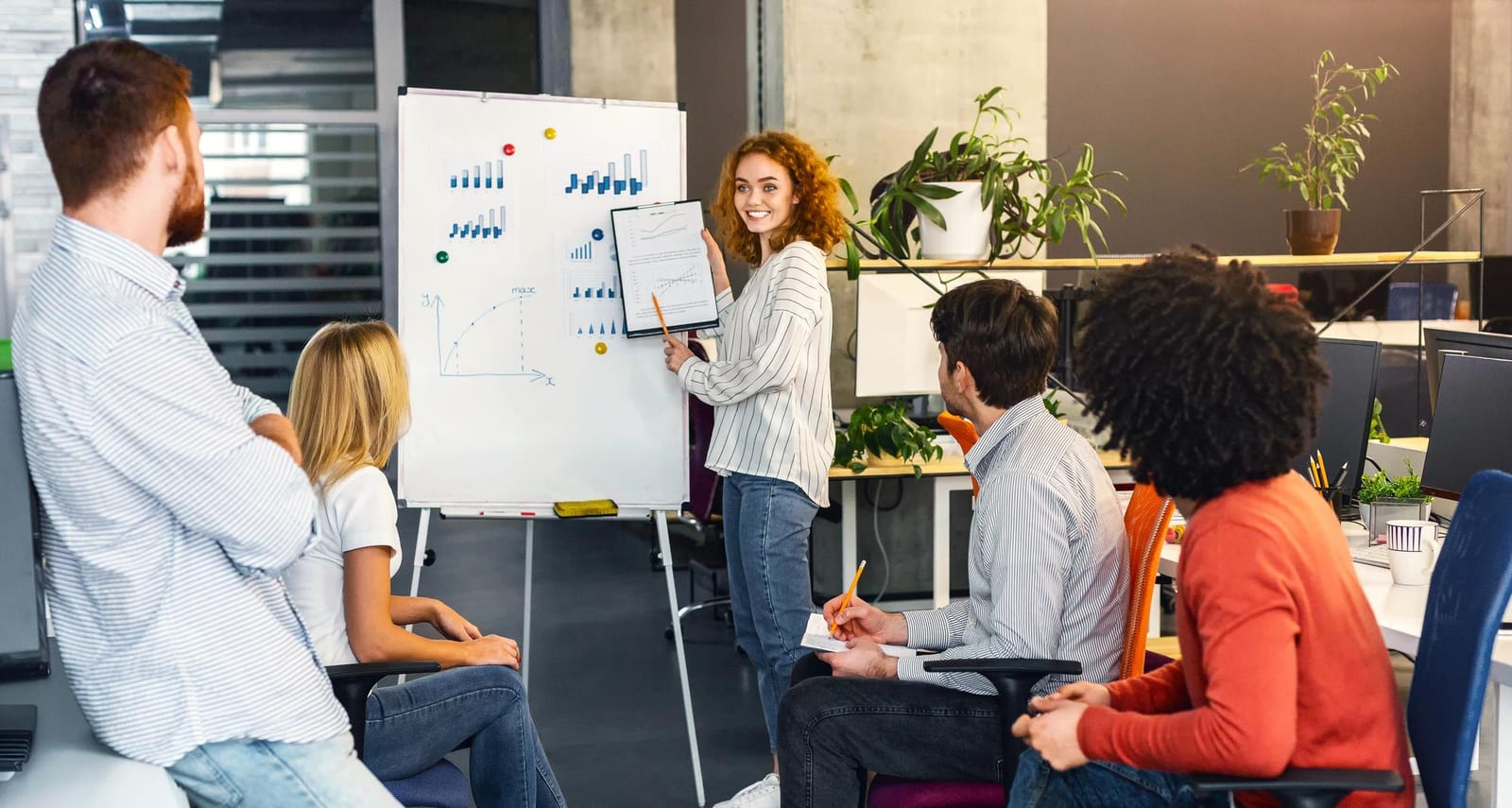 Instrutora aponta para gráficos em um flipchart para colegas sentados em um escritório.